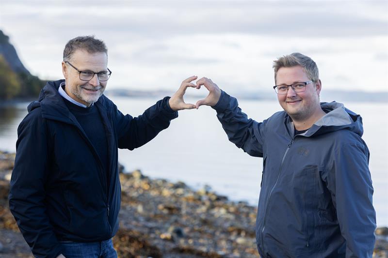 To menn ved fjorden. De lager en hjerteform med hendene.
