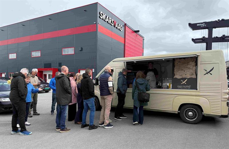 12 personer står i kø for å få lunsj fra foodtrucken til Smaker fra Øyriket. I bakgrunnen ser vi båtbyggeriet til Skarsvaag Boats. Foto: Ida Valsø, Trøndelag fylkeskommune.