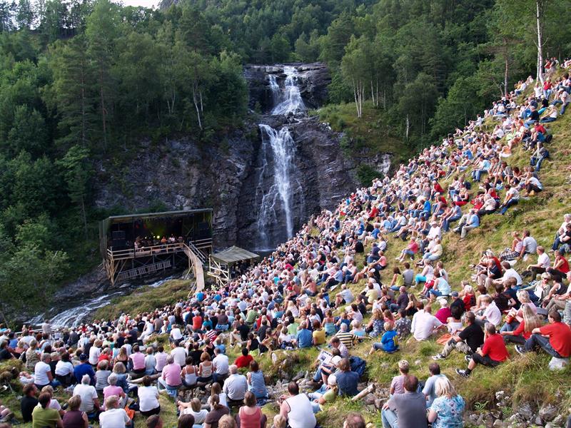 Fosskonserten. Foto, Lars Botten.jpeg Bilde av publikum som sitter i en gresskledd bakke og ser på konsert ved en foss.
