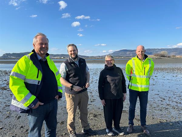 Representanter fra de ulike partene står i fjøra på Hellstranda i Stjørdal. Representantene fra Nye veier har på seg reflekstøy med logo. I bakgrunnen ser vi Trondheimsfjorden speile seg i sola.
