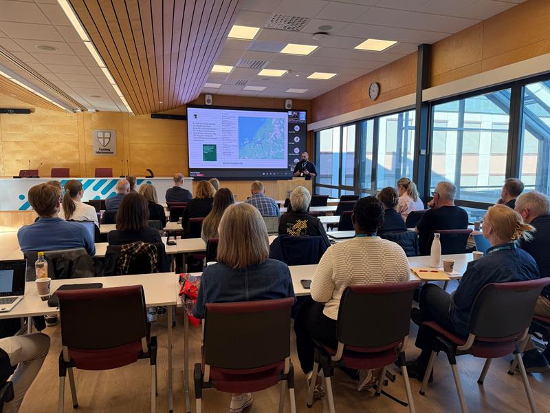 Rundt 30 personer sitter i en sal og ser mot et lerret. Enn mann presenterer.