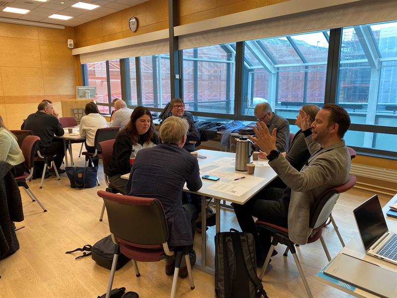 HUNI-3.jpg Seks personer sitter rundt et bord og snakker med hverandre. I bakgrunnen sitter en annen gruppe rundt et bord.
